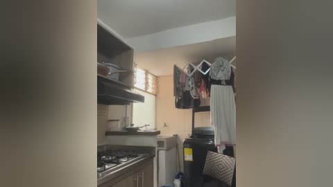 A video of a cluttered, dimly lit kitchen with beige walls, a gas stove, hanging laundry, and a black garbage bin, revealing a chaotic, lived-in space.
