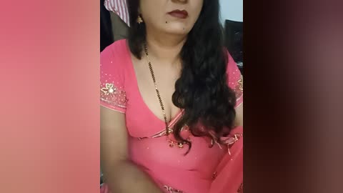 A video of a South Asian woman with medium skin tone, wearing a vibrant pink saree adorned with gold embroidery. She has long, wavy black hair and a red lip.