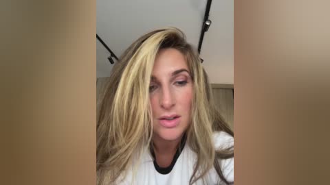 Video of a blonde woman with long, wavy hair, wearing a white top, standing indoors with a wooden background.