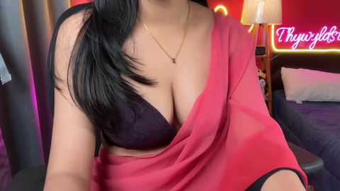 Video of a woman with light brown skin, black hair, wearing a red saree, exposing a black bra, in a dimly lit bedroom with a neon sign and bed.