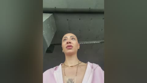 Video of a woman with light skin and dark hair in a bun, wearing a light pink robe and gold necklace, standing in a dimly lit industrial room with exposed metal beams.