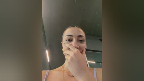 A video of a woman with light skin and dark hair, wearing a white tank top, covering her mouth with her hand, standing in front of a reflective mirror, with blurred, modern background.