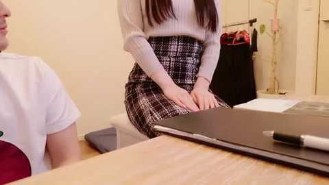 Video of a woman with long dark hair wearing a cream sweater and black-and-white plaid skirt, sitting at a wooden desk with a closed laptop and a pen. Background shows a sewing room with fabric rolls and sewing equipment.
