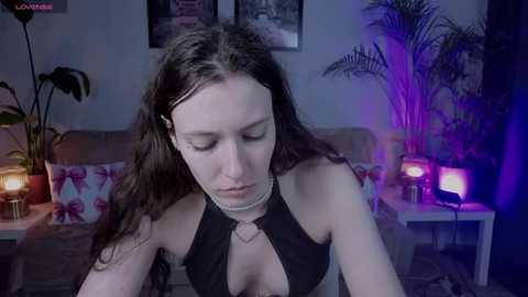 A video of a young woman with long brown hair, wearing a black halter top, sitting in a dimly lit room with purple lighting, a potted plant, and a lamp on a side table.
