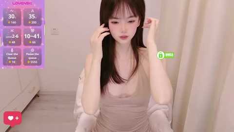Video of an East Asian woman with long black hair, pale skin, and small breasts, wearing a sheer top, adjusting her hair in a minimalist room with a digital clock displaying 10:41.