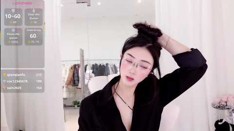 Media: A video of an Asian woman with glasses, fair skin, and black hair, styled in a ponytail, wearing a black coat, in a modern, bright clothing store with racks of clothes visible in the background.