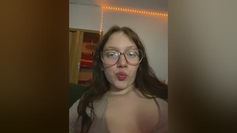 Media: A video of a fair-skinned woman with long brown hair, wearing glasses and a beige top, puckering her lips in a playful manner, indoors with warm lighting and a wooden door in the background.