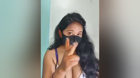 Video of a young South Asian woman with long black hair, wearing a purple bra, partially covered by a black face mask, and pointing her finger in front of her face.