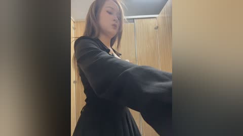 Video of a woman with long brown hair, wearing a black robe, standing in a dimly lit bathroom with beige wooden stalls and a fluorescent light overhead.