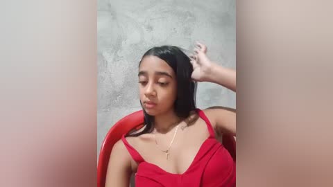 Video of a young woman with medium brown skin, long black hair, and a slender build, wearing a red dress. She sits in a red chair, against a textured gray wall, with one hand in her hair.