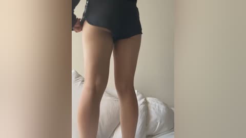 Media: Video of a woman wearing dark shorts, standing in a dimly lit room with beige walls, a bed with white sheets visible in the background.
