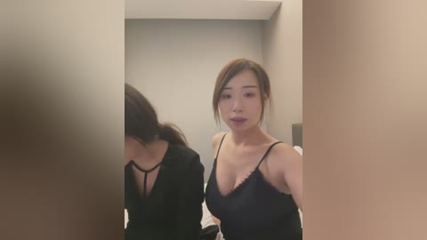 A video of an East Asian woman with long brown hair, wearing a black lace-trimmed spaghetti strap top, standing in a dimly lit, beige-walled room. She has a slender physique and fair skin, with a neutral expression.