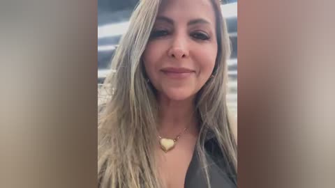 Video of a smiling woman with long, straight, platinum blonde hair, wearing a black jacket and a gold heart necklace. She has a light skin tone and a neutral background with blurred, horizontal lines.