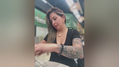A video of a tattooed woman with long brown hair, wearing a black V-neck top, working on a tattoo in a dimly lit tattoo studio.