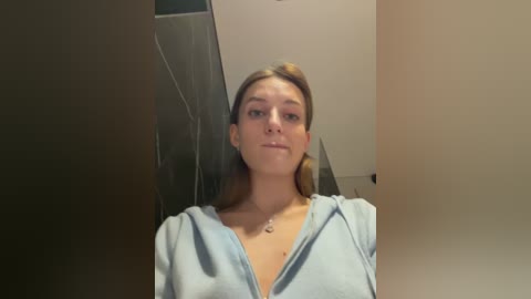 Video of a young Caucasian woman with light skin, straight brown hair, and a light blue hoodie. She stands in a modern bathroom with dark tiled walls and a white ceiling.