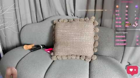 A video of a beige, handmade knit pillow with a scalloped edge on a light grey couch, set against grey curtains. A hand holding a phone with a live stream interface is visible.