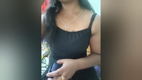 A video of a woman with medium brown skin, wearing a black spaghetti-strap tank top and a delicate gold necklace. She is seated, holding a smartphone. The background is blurred, featuring colorful, patterned fabrics.