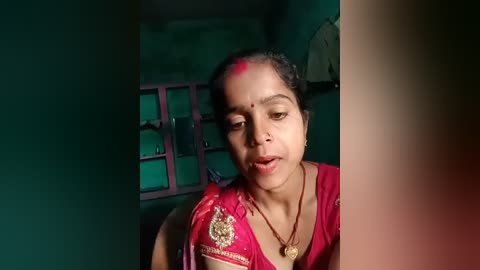 Media: Video of an Indian woman with medium-dark skin, wearing a red sari with gold embroidery, and a gold necklace, applying red powder on her forehead, in a dimly lit room with a green wall and a pink cabinet.