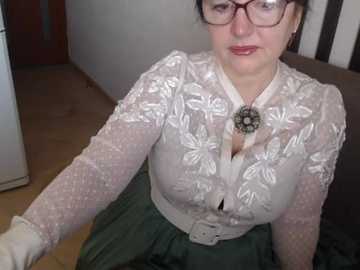 A video of a middle-aged woman with glasses, wearing a lacy white blouse with floral embroidery and a green skirt, sitting indoors with a wooden floor and a white door in the background.