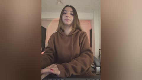 Video of a young woman with light brown skin, straight brown hair, wearing a brown hoodie, sitting in a room with beige walls and a pink door.