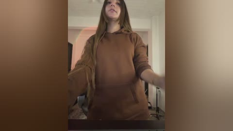 A video of a young woman with long brown hair, wearing a brown hoodie, captured from a low angle in a dimly lit room.