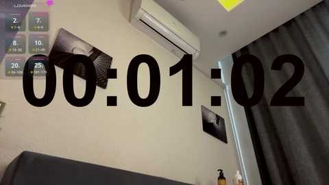 Video of a bedroom with a digital clock reading \"00:01\" on a beige wall. A ceiling-mounted air conditioner and a black framed photo are visible. The room has a grey curtain and a grey sofa.