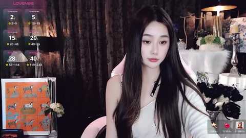 Media: Video of an East Asian woman with long black hair, light skin, and a slender physique, wearing a white top, sitting indoors. Background features dark curtains, a floral arrangement, and a digital display with live stream metrics.