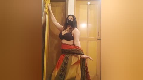 A video of a dark-haired woman with a mask, wearing a black bra and a multicolored sari, leaning against a wooden door in a dimly lit corridor.