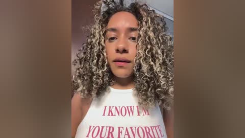 Video of a young woman with light brown skin, curly shoulder-length hair, and a white tank top with red text that reads \"I KNOW YOUR FAVORITE SONG.\