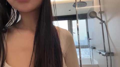 A video of an Asian woman with long, straight black hair, wearing a white tank top, standing in a modern bathroom with a glass shower and metal fixtures.