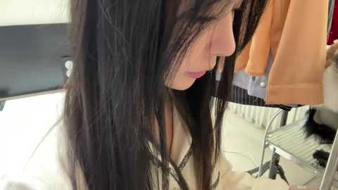 Video of a young woman with long, straight, dark hair, wearing a white top, sitting in front of a mirror, adjusting her hair. Background shows a black headboard, a white radiator, and a peach-colored garment hanging on a hook.