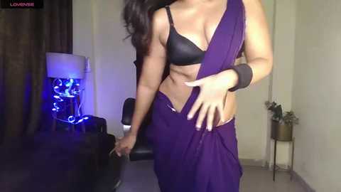 Media: Video of a curvy woman in a purple sari, black bra, and wrist cuffs, standing in a dimly lit room with a blue-lit bookshelf and a potted plant.