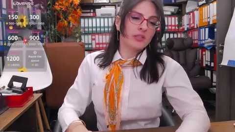 Media: Video of a woman with glasses, wearing a white shirt and orange tie, seated at a desk in a colorful, organized office.