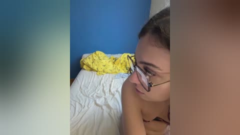 Video of a young woman with glasses lying on a bed, wearing a yellow-patterned bra. Her face is turned slightly to the side, revealing a calm expression. The background features a blue wall and white bedding.