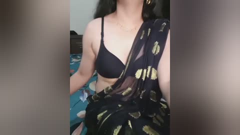 Media: Video of a woman with light brown skin, wearing a black bra and a black sari with gold leaf patterns, sitting on a blue-patterned bed.