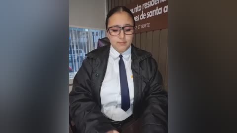 Video of a young Latina woman with fair skin, glasses, dark hair, and a white shirt with a navy tie, sitting at a desk in a library.