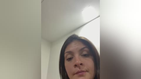 A video of a young woman with straight brown hair, fair skin, and a neutral expression, captured from a low angle in a white-walled room with a ceiling light visible.