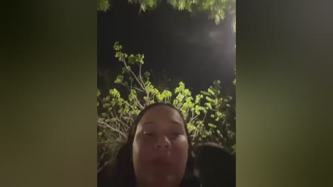 Video of a serene Asian woman with closed eyes, positioned centrally in a dark, outdoor setting with green foliage and a faint light source above.