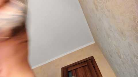 A video of a beige-tiled bathroom ceiling with a textured wall, featuring a blurry person with light skin and a blonde ponytail, wearing a dark bra. The image captures a close-up, low-angle perspective.