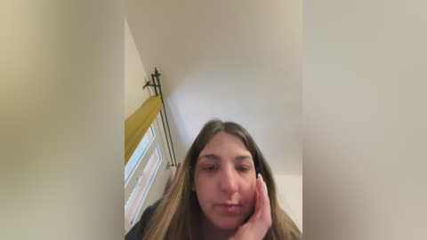 Media: Video of a young woman with long, light brown hair, resting her face on her hand in a casual indoor setting. The room has a white ceiling and a yellow window blind.