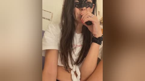 Media: Video of a woman with long dark hair, wearing a white cropped top, black lace mask, and a black scrunchie, holding a phone to her mouth, against a beige wall.