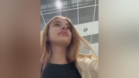 Media: A video of a young, fair-skinned woman with long, straight blonde hair, wearing a black top and beige jacket, gazing upward in a modern, dimly lit room with white ceiling tiles and recessed lighting.