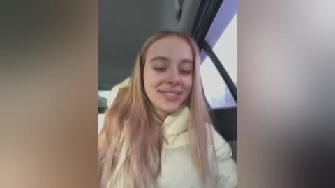 Media: Video of a young Caucasian woman with long, straight blonde hair and fair skin, wearing a light-colored puffer jacket, smiling. Background shows a car interior with tinted windows and blurred urban landscape.