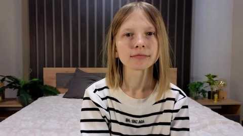 Video of a young Caucasian girl with straight, shoulder-length blonde hair, wearing a black and white striped shirt, standing in a modern bedroom with a gray bed, wooden headboard, and vertical striped curtains.