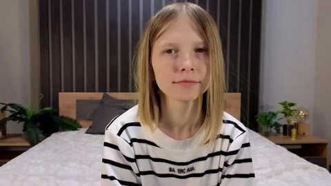 Video of a blonde girl with straight hair, wearing a black and white striped shirt, sitting on a bed in a modern bedroom with grey curtains and green plants.