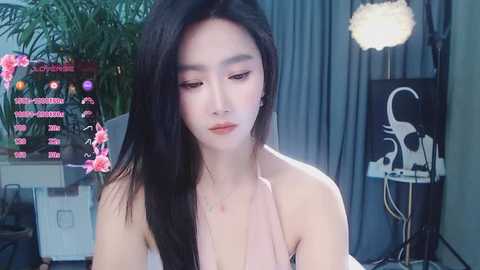 Media: Video of a young East Asian woman with long black hair, wearing a pink dress, standing in a modern, well-lit room with a potted plant, wall art, and a spotlight.