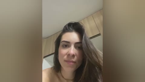 Video of a young woman with medium skin tone and long, dark hair, wearing a tan top, standing in a room with beige walls and light wooden cabinetry.