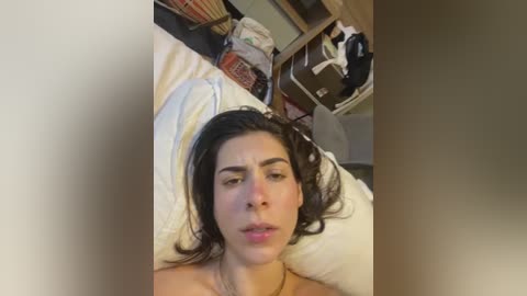 Video of a young woman with olive skin, dark hair, and expressive eyes, lying on a bed, surrounded by a cluttered bedroom with wooden furniture and a fan.