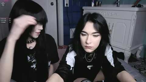 Video of a young woman with pale skin and long black hair, wearing a black shirt with white accents, sitting on a bed. She has a gothic style with dark makeup and a spiked collar. Background shows a white dresser and a mirror.