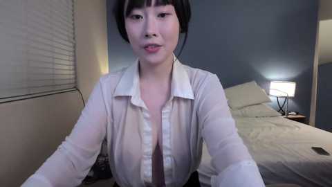 A video of an Asian woman with short black hair, wearing a see-through white blouse, sitting on a bed with a white blanket. The room has a blue wall and a lit lamp.